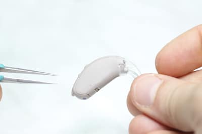 Regular Hearing Aid Servicing and Repairs Audiologist Performs Hearing Aid Servicing image is a closeup showing fingertips holding a hearing aid on the right while a pair of tweezers is in the frame on the left.