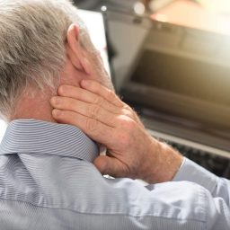 Older man looking down at a computer has his hand on that back of his neck that is in pain.
