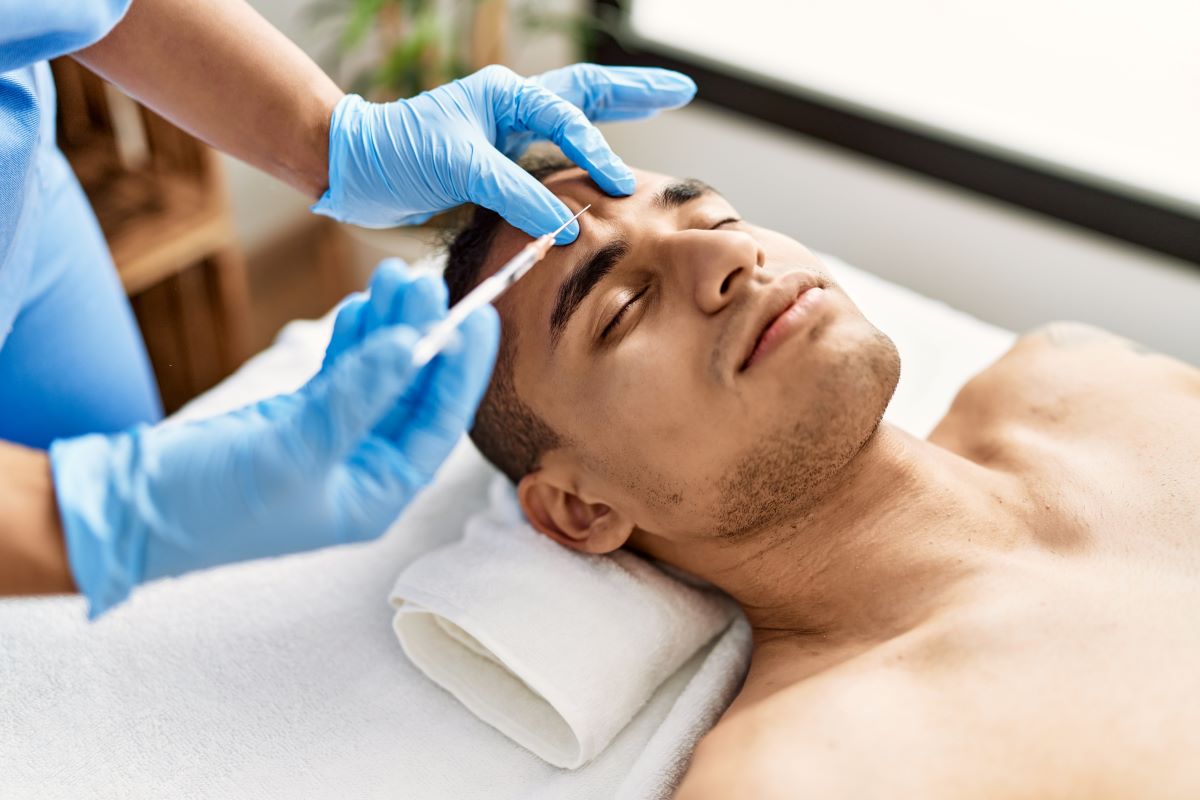 Man in a semi-clinical setting laying down on a table with a towel under his neck. Photo is just of mans shoulders and face. A pair of blue gloved hands is squeezing above his eyebrow in order to inject a solution.